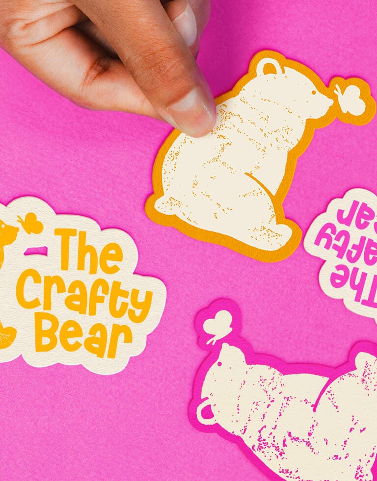 A close-up of a hand placing a sticker of the bear-and-butterfly logo on a pink fabric surface. Several variations of "The Crafty Bear" stickers are scattered around, including yellow-on-white, white-on-orange, and purple-pink combinations. The stickers are die-cut and playful, emphasizing a fun, handmade brand aesthetic.
